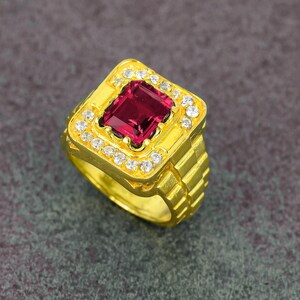 May include: A gold-colored ring with a large, square, deep red gemstone at the center. The gemstone is surrounded by smaller, clear stones. The ring has a textured band and a square face.