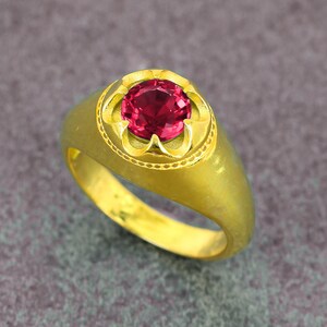May include: A gold ring with a large, round, red gemstone. The gemstone is set in a decorative bezel. The ring has a smooth, polished band.