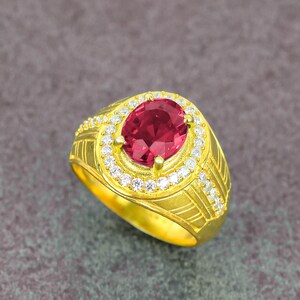 May include: A gold-colored ring with an oval, deep red gemstone at the center, surrounded by a halo of smaller, clear stones. The ring's band features etched linear designs and additional small stones.