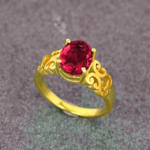 May include: A gold ring with a large, oval-cut, deep red gemstone. The ring has an ornate, filigree design on the band and around the gemstone setting. The ring is displayed on a grey surface.