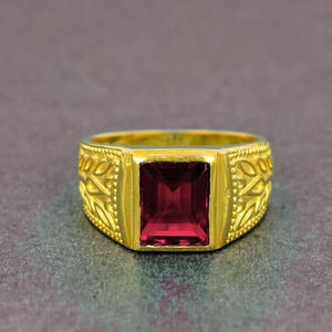 Può includere: Un anello d'oro con una pietra preziosa rosso scuro rettangolare. L'anello ha un design ornato sulla fascia con un motivo strutturato. La pietra preziosa è il punto focale, incastonata in una lunetta d'oro.