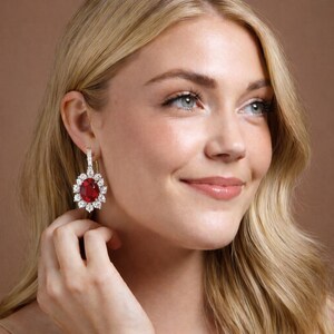May include: A close-up of a woman wearing a pair of elegant earrings. Each earring features a large, oval, deep red gemstone surrounded by a halo of smaller, sparkling white stones. The earrings are set in a gold-colored metal and dangle from a small hoop.