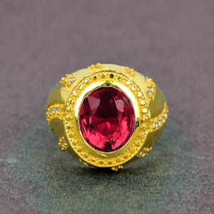 May include: A gold-colored ring featuring a large, oval-shaped, ruby-red gemstone. The ring has a textured band with small, clear accent stones and decorative detailing around the central stone. The ring is set against a dark gray background.