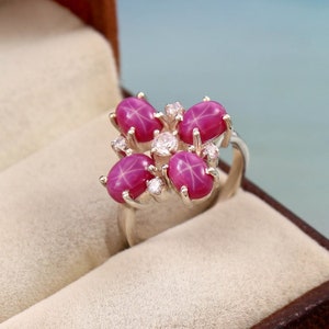 May include: A silver ring with five oval, star-shaped, deep pink gemstones arranged in a cluster. Smaller, clear gemstones are set between the larger stones. The ring is displayed against a neutral background, highlighting the jewellery's design.