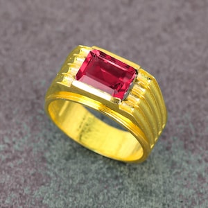 May include: A gold ring featuring a rectangular, deep red gemstone. The ring has a ribbed design on the sides and a polished, shiny finish. The gemstone is set in a gold bezel.