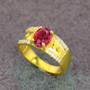May include: A gold ring featuring a large, oval-cut, deep red gemstone. The ring's band is also gold, with small, clear gemstones set along the sides. The ring is a piece of jewelry.