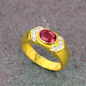 May include: A gold ring with a large oval ruby center stone. The ring is accented with small, round, white gemstones on each side of the ruby. The ring has a thick band and a polished finish.