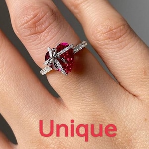 Könnte beinhalten: Ein silberner Ring mit einem herzförmigen, tiefroten Edelstein. Der Edelstein ist mit einer silbernen Schleife verziert und mit kleinen, klaren Steinen entlang des Bandes besetzt. Das Wort "Unique" wird in rotem Text angezeigt.