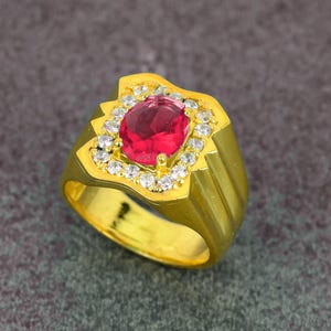 Puede incluir: Un anillo dorado con una gran gema roja ovalada en el centro, rodeada de piedras más pequeñas y transparentes. La banda del anillo tiene un diseño texturizado. El anillo está sobre un fondo gris.