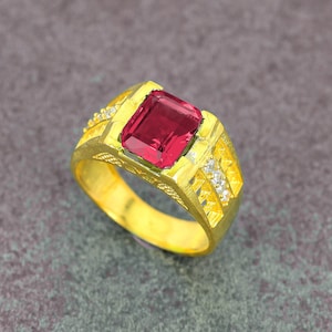 May include: A gold ring featuring a large, rectangular, red gemstone at the center. The ring has a detailed design with small, clear accent stones and intricate patterns along the band. The ring is set against a gray background.