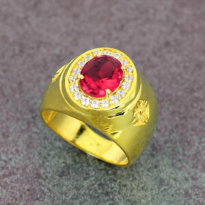 Peut inclure: Bague dorée ornée d'une grosse pierre rouge ovale, entourée de petits brillants. La bague présente un anneau épais et des détails décoratifs sur les côtés. Un bijou classique.