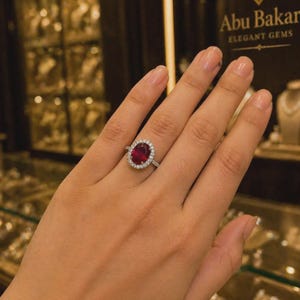 Peut inclure: Une main présente une bague en argent ornée d'un grand rubis ovale, entouré d'un halo de petites pierres claires. La bague est mise en valeur sur un fond d'écrans de bijoux. Le texte "Abu Bakar Elegant Gems" est visible.