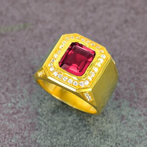 May include: A gold-colored ring with a large, rectangular, deep red gemstone at the center. The gemstone is surrounded by smaller, clear stones. The ring has a thick band and a square face, showcasing a classic, luxurious design.