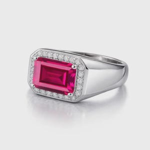 Puede incluir: Un anillo plateado con una gran gema rosa brillante de forma rectangular. La gema está rodeada por un halo de pequeñas piedras claras. El anillo tiene una banda pulida y lisa.