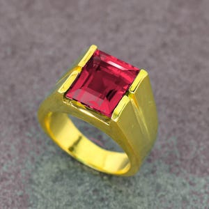 May include: A gold ring featuring a large, square-cut, deep red gemstone. The ring has a thick, polished gold band with a classic, masculine design. The gemstone is the focal point, catching the light and showcasing its rich color.