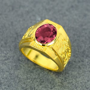 Può includere: Un anello d'oro con una gemma ovale rosso scuro. L'anello presenta un design ornato e inciso sui lati e una finitura lucida e brillante. La gemma è incastonata in una montatura a castone.