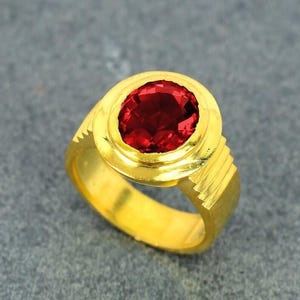 Puede incluir: Un anillo de oro con una gran gema roja oscura de corte ovalado. El anillo tiene una banda gruesa y un engaste detallado alrededor de la piedra. El oro tiene un acabado pulido.
