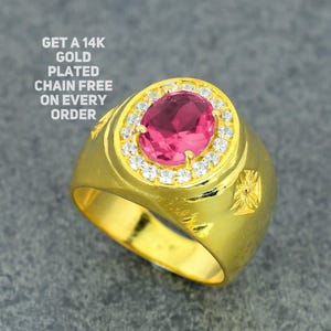 Pode incluir: Um anel banhado a ouro com uma grande pedra preciosa rosa oval no centro, rodeada por pedras menores e transparentes. O anel tem uma banda larga e elementos decorativos nas laterais. Texto na imagem: GET A 14K GOLD PLATED CHAIN FREE ON EVERY ORDER.