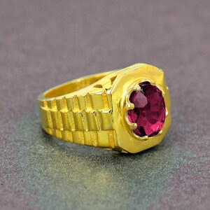 Peut inclure: Une bague dorée avec une grosse pierre précieuse rouge rubis de forme ovale. La bague a un anneau texturé et un sertissage carré autour de la pierre précieuse. La bague est posée sur une surface grise.