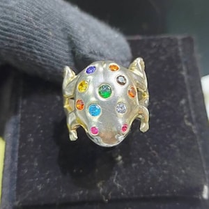 Puede incluir: Un anillo de plata con un diseño de rana detallado. La rana está adornada con piedras preciosas de colores, incluyendo rojo, naranja, amarillo, verde, azul y morado.