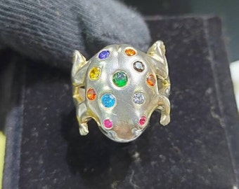 May include: A silver ring with a detailed frog design. The frog is adorned with colorful gemstones, including red, orange, yellow, green, blue, and purple.