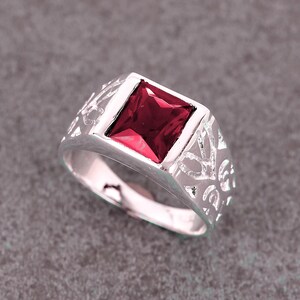 Può includere: Un anello d'argento con una gemma quadrata rosso scuro. La fascia presenta un design ornato e inciso. La gemma è incastonata in una lunetta rettangolare. L'anello è mostrato su una superficie grigia.