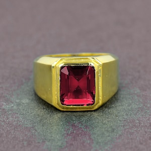 May include: A gold-colored ring with a large, rectangular, deep red gemstone. The ring has a thick band and a simple, classic design. The gemstone is set in a square bezel.