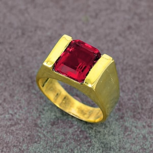 Könnte beinhalten: Ein goldener Ring mit einem großen, rechteckigen, tiefroten Edelstein. Der Ring hat ein dickes Band und eine Fassung, die den Edelstein sicher hält. Der Ring wird auf einem neutralen Hintergrund präsentiert.