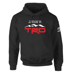 May include: A black hooded sweatshirt featuring a white mountain graphic above the red text TRD and the white text TOYOTA RACING DEVELOPMENT. The sleeve displays a white Toyota logo and the word TOYOTA. The garment includes a front kangaroo pocket.