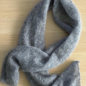 May include: A soft, gray scarf made of a fuzzy material. The scarf is draped on a light-colored wooden surface. The scarf is a solid color and appears to be lightweight, suitable for cooler weather. The scarf is a simple design.