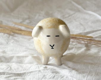 以下が含まれることがあります： クリーム色の体と白い顔の陶器製の羊の置物。羊は小さな黒い目と黒い「Y」字型の口をしています。置物は4本の小さな脚の上に立っており、2つの小さな耳があります。羊は白い布の上にあります。