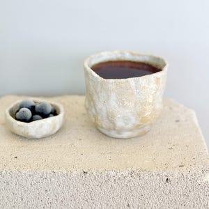 Könnte beinhalten: Eine strukturierte, cremefarbene Keramiktasse, gefüllt mit einer dunklen Flüssigkeit, möglicherweise Tee oder Kaffee, steht neben einer kleinen Schale mit dunklen Beeren. Die Tasse und die Schale stehen auf einer hellen, strukturierten Oberfläche, was eine minimalistische Ästhetik erzeugt.