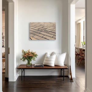 May include: A hallway scene featuring a dark wood bench with black metal legs, two white pillows, and a vase of flowers. A framed artwork with text is mounted on the white wall. The floor is dark wood.