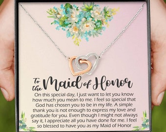 Puede incluir: Un collar plateado con dos colgantes de corazón entrelazados, uno de oro rosa y otro plateado, presentado en una caja de regalo. La caja presenta un diseño floral y el texto "To the Maid of Honor", con un mensaje sincero.