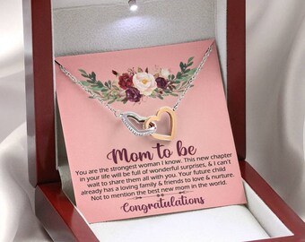 Puede incluir: Una caja de madera con una tarjeta rosa en su interior. La tarjeta tiene un diseño floral y el texto "Mom to be". La tarjeta va acompañada de un collar de plata y oro con dos corazones entrelazados.