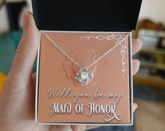 Puede incluir: Un collar de plata con un colgante de nudo y una piedra transparente en el centro. El collar está en una caja con fondo rosa y el texto "¿Quieres ser mi dama de honor?"