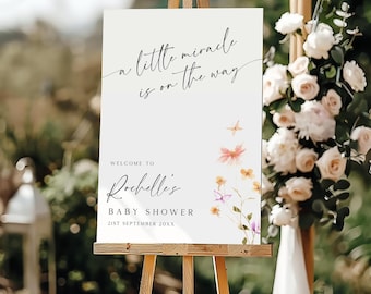 Op de afbeelding: Een wit bord met een bloemenontwerp en de tekst "a little miracle is on the way". Het bord bevat ook de tekst "Welcome to Rochelle's Baby Shower 21st September 20XX".