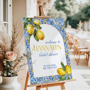 May include: Welcome to Hannah's bridal shower sign with a blue and white tiled border and lemons. The sign says "Welcome to Hannah's bridal shower" and "42 days until she says I do!"