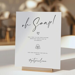 Peut inclure: Un panneau blanc avec le texte "oh Snap!" en écriture noire élégante, un symbole de cœur et des instructions liées à Snapchat. Le panneau est maintenu par un support en métal doré, adapté à un mariage ou une fête.