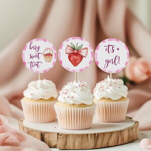 Könnte beinhalten: Drei Cupcakes mit weißem Zuckerguss und rosa Streuseln, gekrönt von dekorativen Aufsätzen. Die Aufsätze zeigen eine Erdbeere, einen Korb mit Erdbeeren und den Text "It's a girl!" auf einem rosa-weiß karierten Hintergrund. Die Cupcakes stehen auf einem Holzsockel.