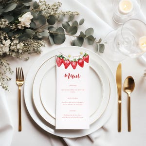 Könnte beinhalten: Eine formelle Tischdekoration mit einem weißen Teller, goldenem Besteck und einer Menükarte. Die Menükarte zeigt eine Aquarellillustration von Erdbeeren und Gänseblümchen mit dem Wort "Menu" in roter Schrift. Der Tisch ist mit Grün und Kerzen dekoriert.