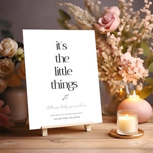 May include: A white sign with the text "it's the little things" in black, displayed on a small wooden easel. The sign includes the text "Please help yourselves" and the names "JACOB AND HELAENA". The background features flowers and candles.