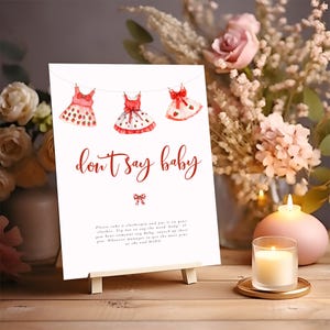 Könnte beinhalten: Ein weißes Schild mit dem Text "don't say baby" in roter Schrift, mit Aquarellillustrationen von Babykleidern. Das Schild wird auf einer kleinen Holzstaffelei präsentiert, mit einer Kerze und einem Blumengesteck im Hintergrund.