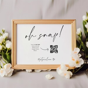 Peut inclure: Un panneau encadré doré avec l'inscription "oh snap!" en écriture cursive. Le panneau comprend un code QR et le texte "Scannez le code QR ci-dessous et ajoutez des photos à notre album Google Photos." Des fleurs blanches ornent le cadre.