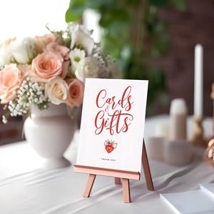 Könnte beinhalten: Ein weißes Schild auf einer kleinen Staffelei mit den Worten "Cards & Gifts" in roter Schrift und einer Erdbeerillustration. Das Schild steht auf einer weißen Tischdecke, mit einem Blumenarrangement und Kerzen im Hintergrund. Der Text "THANK YOU!" steht unten.