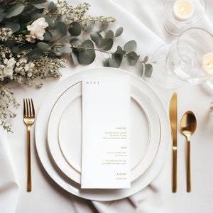 Puede incluir: Un menú de boda blanco con letras doradas sobre una mesa blanca con cubiertos dorados y flores blancas. El menú incluye entrantes, plato principal y postre.