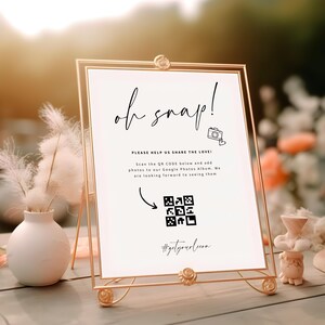 Peut inclure: Un panneau doré avec l'inscription "oh snap!" en écriture élégante. Le panneau comprend un code QR et le texte "PLEASE HELP US SHARE THE LOVE!" et "#getyourloom". Un petit vase blanc avec des plumes décoratives et des fleurs est en arrière-plan.