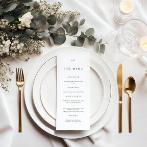 May include: Elegant table setting with white plates, gold cutlery, and a menu card. The menu card reads "The Menu" with initials at the top. Floral arrangement and candles add to the sophisticated ambiance. Perfect for a wedding or formal event.