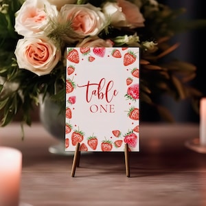 Könnte beinhalten: Eine Tischkarte mit den Worten "Table One" in roter Schrift, umgeben von Aquarell-Erdbeeren. Die Karte wird von einer kleinen Holzstaffelei gehalten. Ein Strauß rosa Rosen und brennende Kerzen befinden sich im Hintergrund.