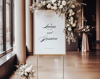 May include: A gold-framed wedding welcome sign with the names "Lucas and Jessica" in script. The sign is decorated with white flowers and foliage, and the date "02 MAY 2023" is shown. A floral arrangement is at the base. The sign is on a gold stand.